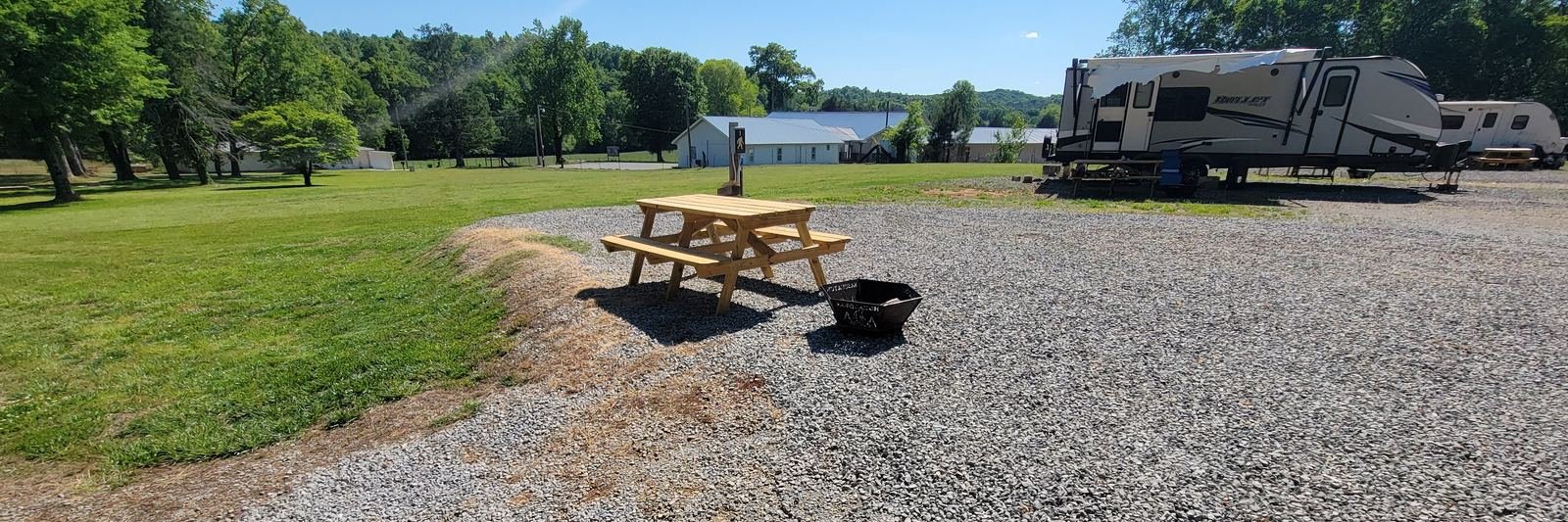 A wooden picnic table and steel fire pit on a gravel site at Gryptid RV Park, with a travel trailer at the next site